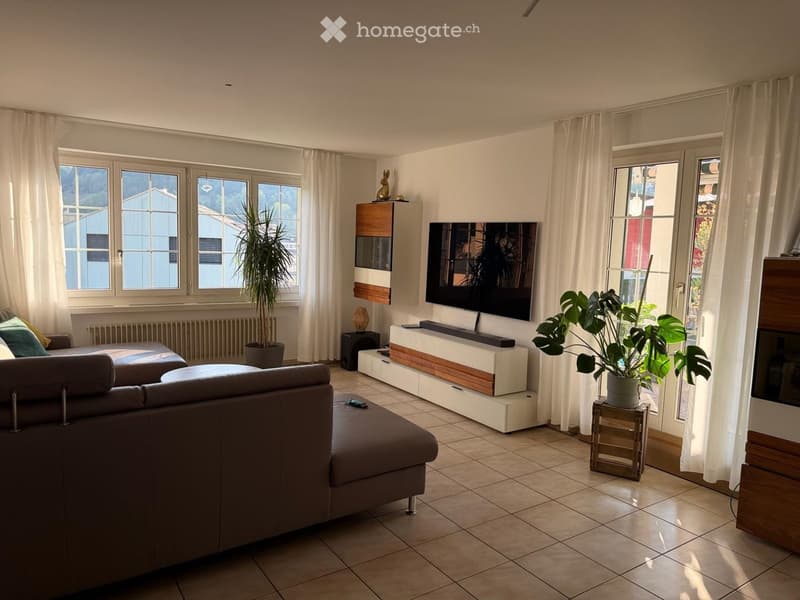 Modern living room with a brown leather sofa, ottoman, coffee table, flat-screen TV, side table with a plant, and a cabinet with a decorative item on top.