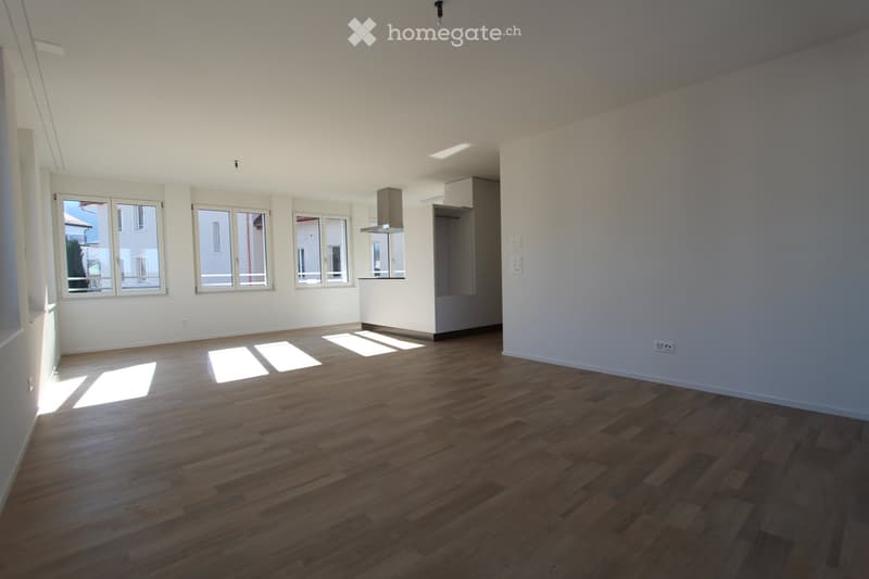 Empty room with wooden flooring, white walls, and large windows allowing natural light to enter. The room is unfurnished and features a small kitchen area with a built-in cabinet and a range hood visible in the background.