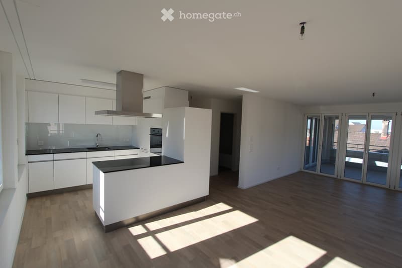 Modern, unfurnished kitchen with white cabinets, a central island, and large windows leading to a balcony. The space features wooden flooring and a sleek, minimalist design.