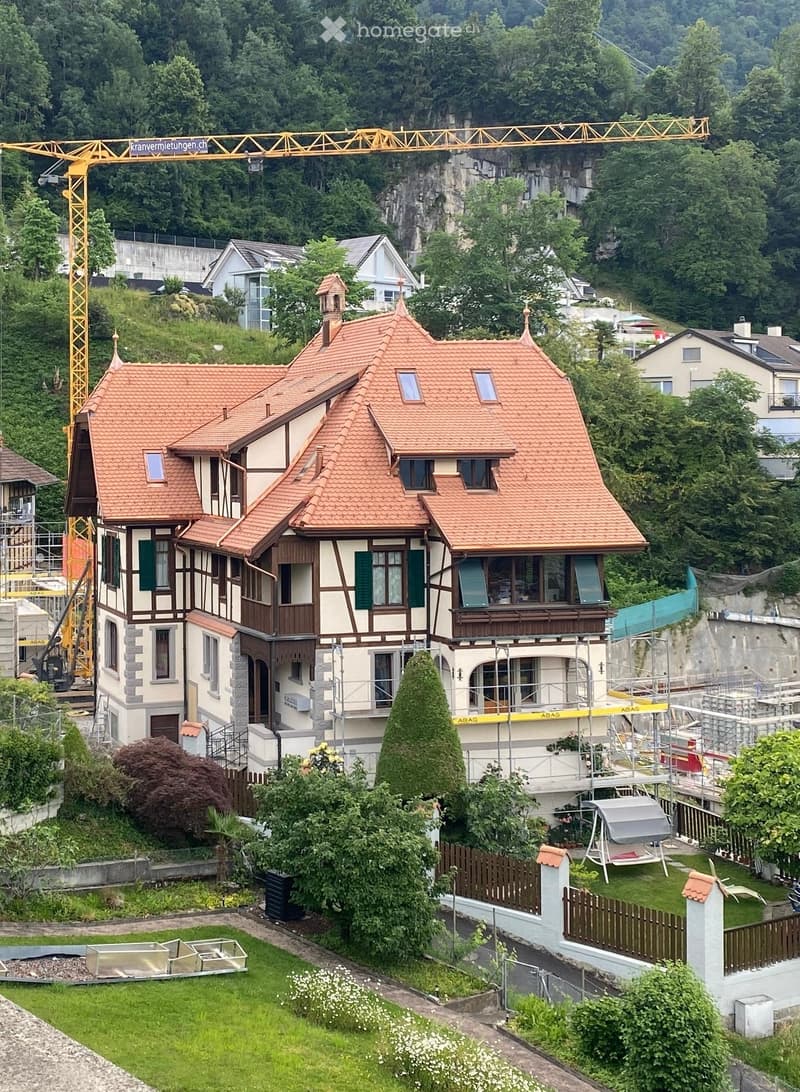 A multi-story house with a red-tiled roof, green shutters, and a balcony, surrounded by a well-maintained garden and trees. A crane is visible in the background, indicating construction or renovation work.
