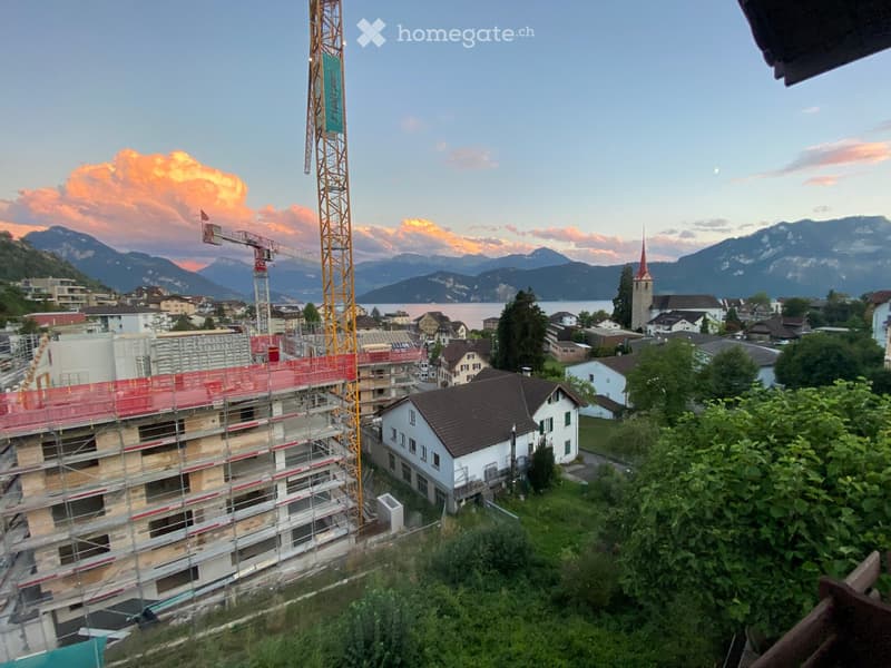 View of a town with a partially constructed building covered in scaffolding, a tall crane, and a church tower in the background, with mountains and a lake visible in the distance.