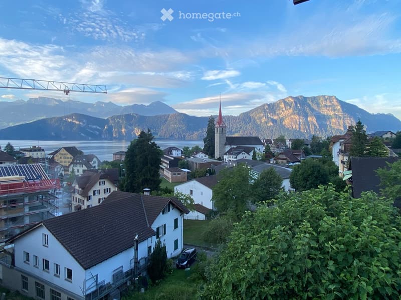 A scenic view of a town with houses, a church tower, and mountains in the background, with a body of water visible in the distance.