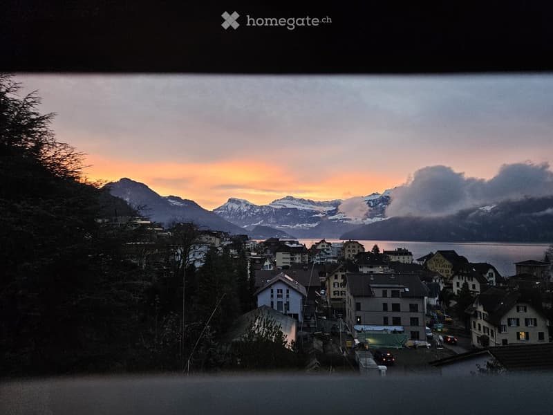A scenic view of a town nestled in a valley with snow-capped mountains in the background, featuring multiple houses and buildings, a lake, and a sunset sky.