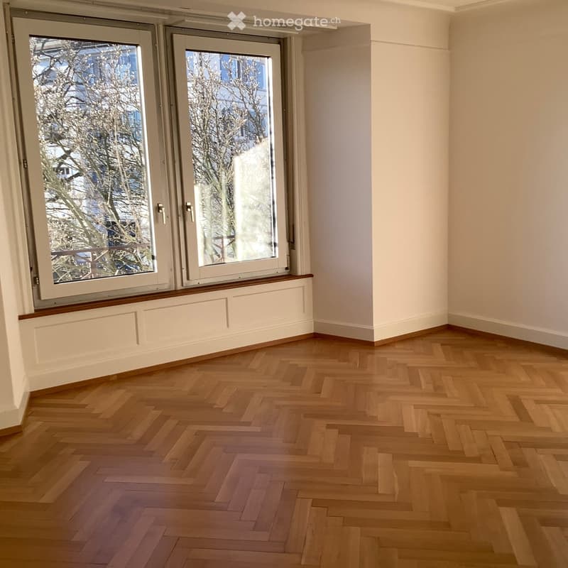 Empty room with wooden herringbone flooring, white walls, and large windows with white frames and a radiator cover below them.