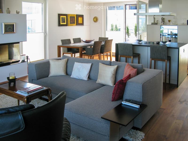 Modern living room with a grey sectional sofa, coffee table, dining area with chairs and table, kitchen island with bar stools, and decorative items including candles and artwork.
