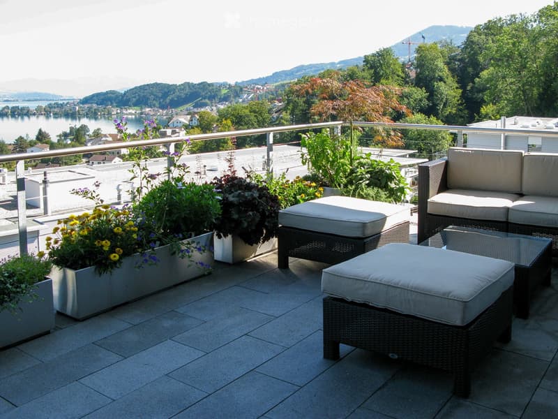 Balcony with wicker sofa, ottomans, and a small table, surrounded by potted plants, overlooking a scenic lake and mountains.