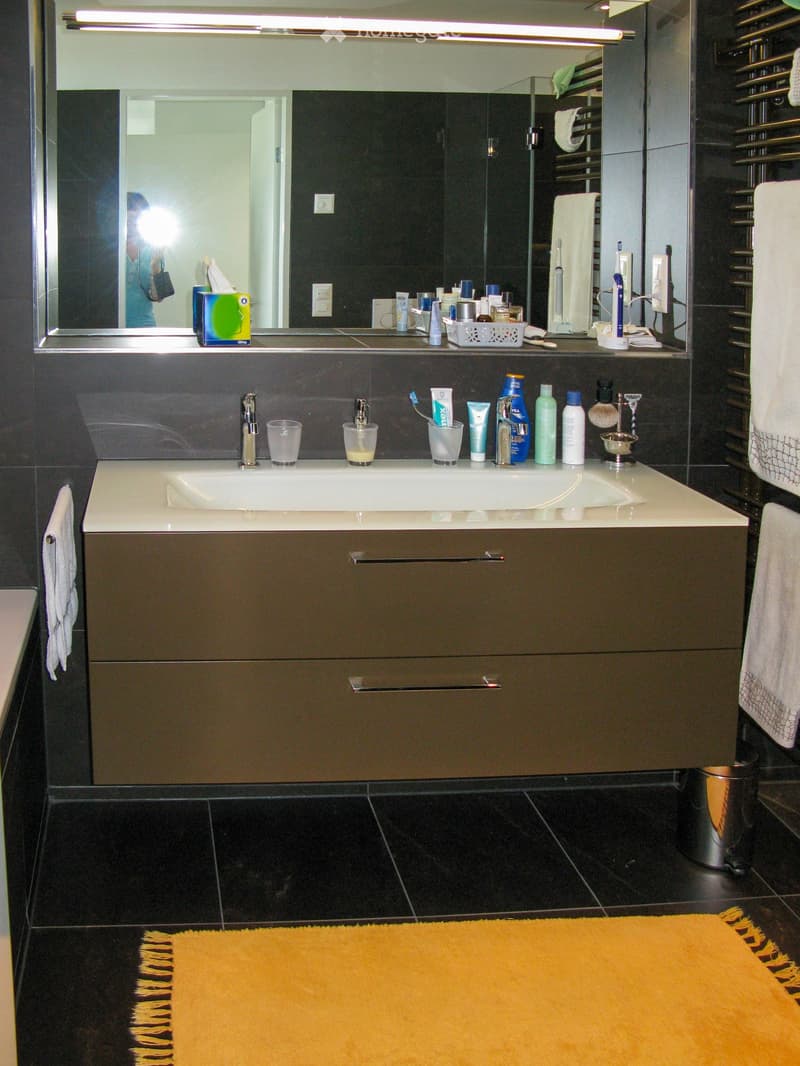 Modern bathroom with a double sink vanity, mirror, various toiletries, and a yellow bath mat on the floor.