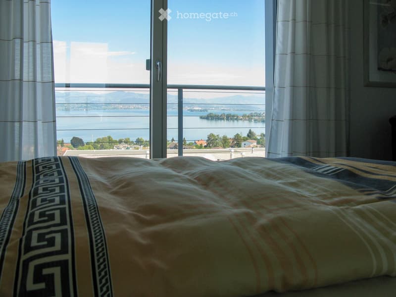 Bedroom with a large window offering a scenic view of a lake and mountains, featuring a bed with patterned bedding.