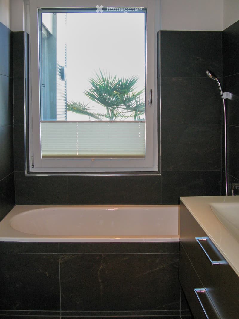 Modern bathroom with a bathtub, sink, and large window overlooking a palm tree. Dark tiled walls and floor, sleek fixtures.