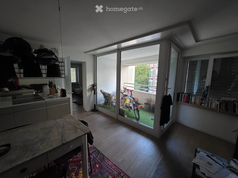 Bright living space with wooden flooring, large sliding glass doors opening to a balcony, a kitchen area with cabinets and a pendant light, a dining table with chairs, a bookshelf, and a bicycle on the balcony.