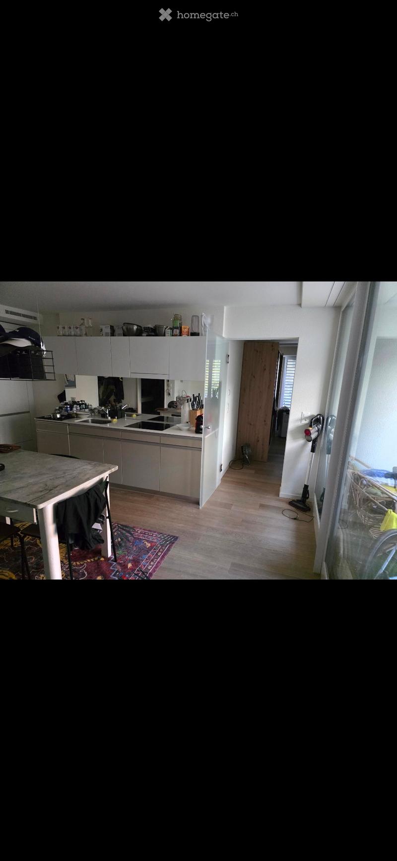 Modern kitchen with grey cabinets, a central island, and a dining area with a table and stool. The floor is wooden, and a colorful rug is placed near the island.