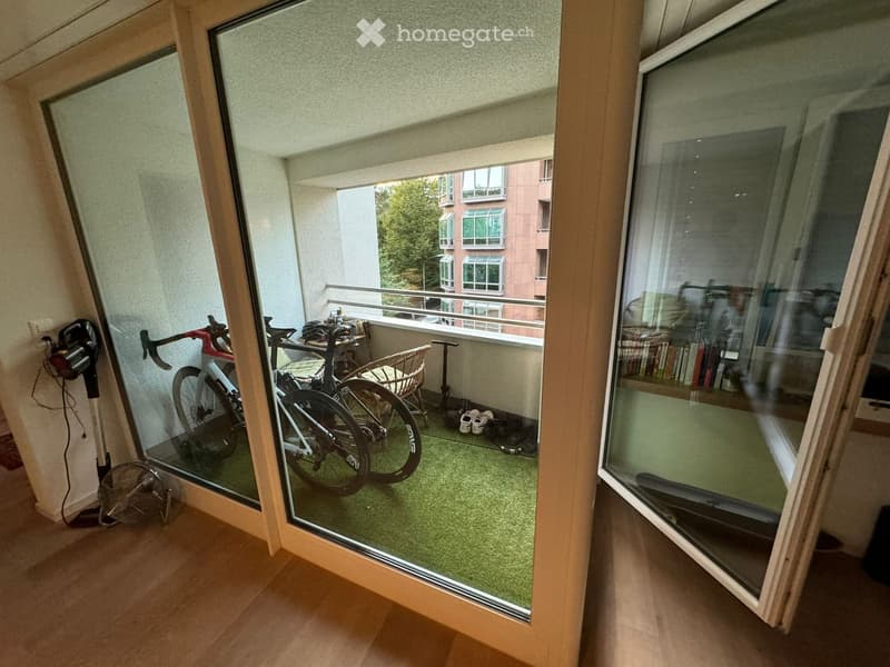 Balcony with green artificial turf, two bicycles, a small table, a chair, and a fan. The balcony has large glass doors leading to the interior.