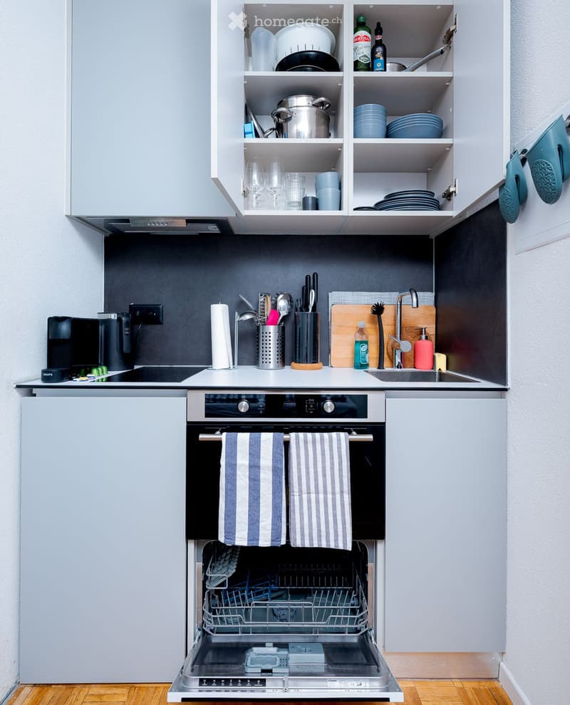 Open dishwasher, silver oven, black countertop, stainless steel sink, modern appliances, white cabinets, dark backsplash