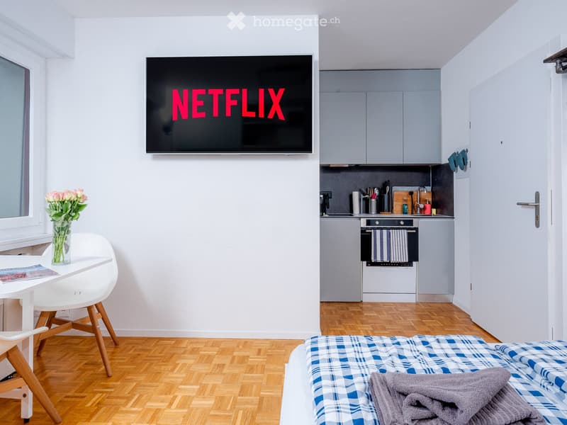 Small room, wooden floors, white walls, bed, small kitchen, tv on wall, table and chair