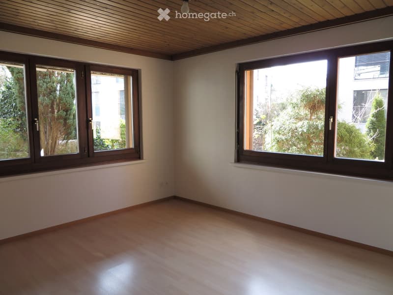 Empty room with wooden floor, white walls, and two large windows with wooden frames. View of trees outside.