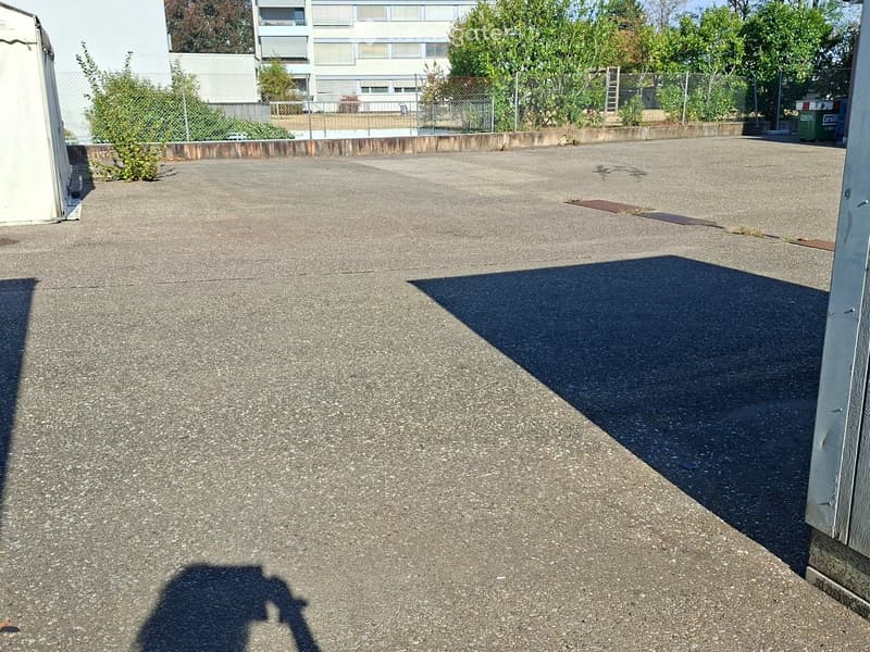 Empty rooftop terrace with concrete surface, surrounded by a low fence and greenery, with a building in the background.