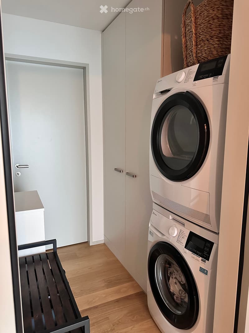 A small, modern laundry room with a stacked washer and dryer, a wooden bench, and white cabinetry.