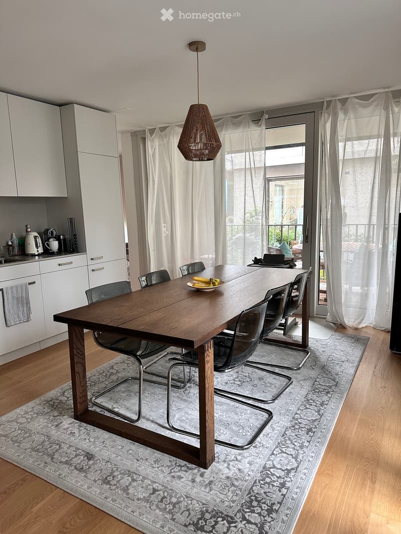 Modern dining area with a large wooden dining table surrounded by chairs, a kitchen area with white cabinets and appliances, and large windows with sheer curtains leading to a balcony.
