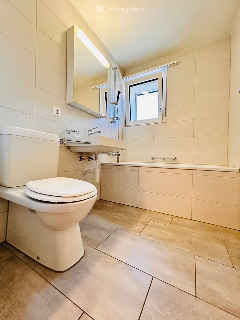 Modern bathroom with white tiles, a toilet, a sink, a bathtub, and a mirror above the sink. The room features a window with a frosted glass design and a sleek, contemporary aesthetic.