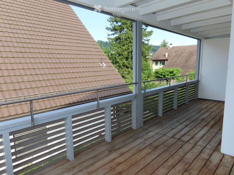 Wooden balcony with white railings, overlooking a tiled roof and surrounding greenery.