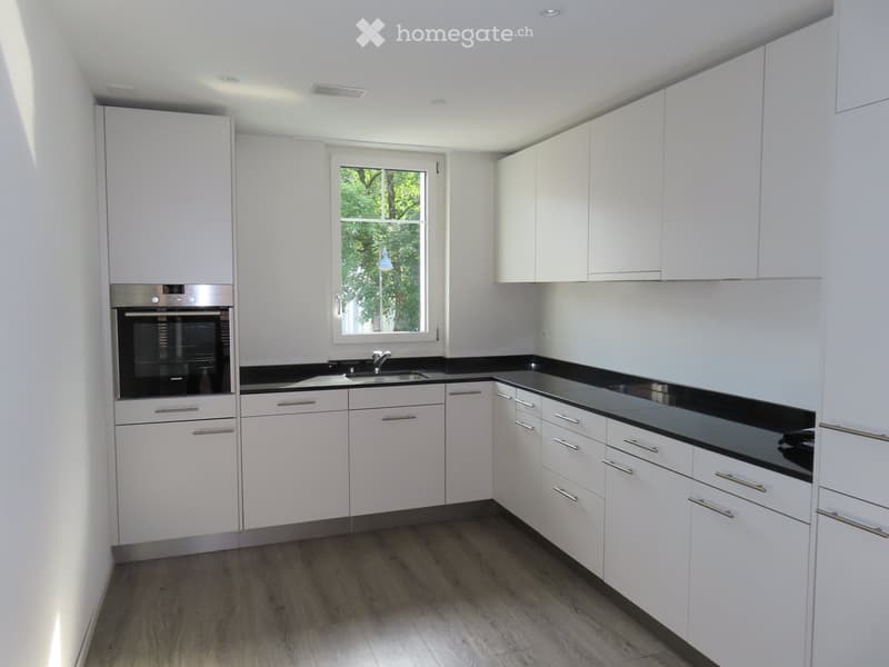 Modern kitchen with white cabinets, black countertops, and a stainless steel oven. Features a window with a view of greenery outside.