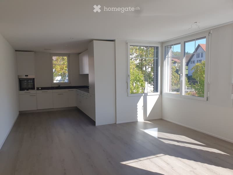 Empty kitchen with white cabinets, dark countertops, and large windows letting in natural light. The floor is light wood.