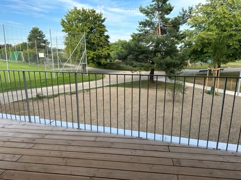 Wooden balcony with metal railing, overlooking a green park area with trees and pathways.