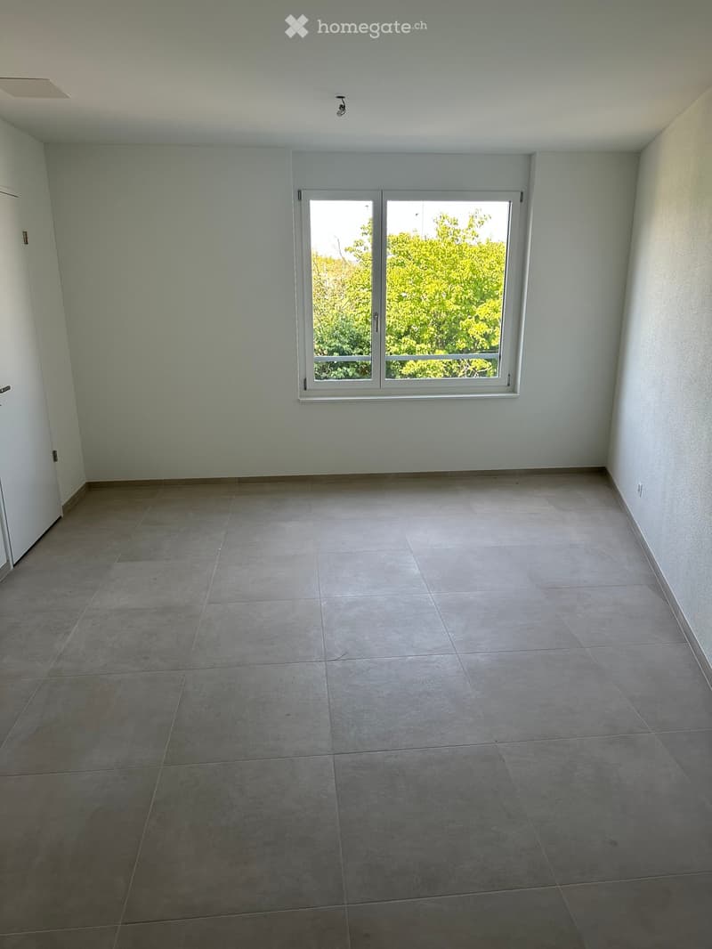Empty room with tiled floor, white walls, and a large window with a view of greenery outside.