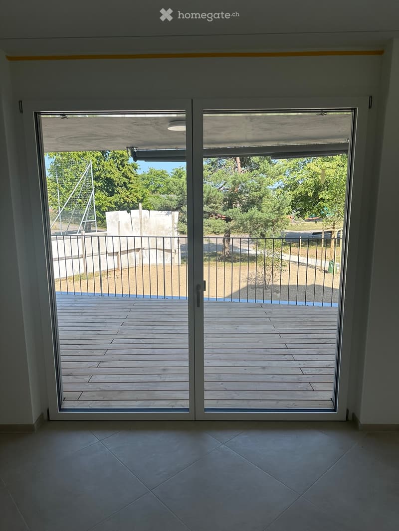 Modern interior with large sliding glass doors opening to a wooden decked balcony area, featuring a metal railing and outdoor greenery visible beyond.