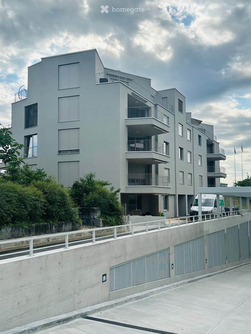Modern multi-story residential building with grey facade, multiple balconies, and large windows. Features a raised ground floor with a concrete barrier and parking area.