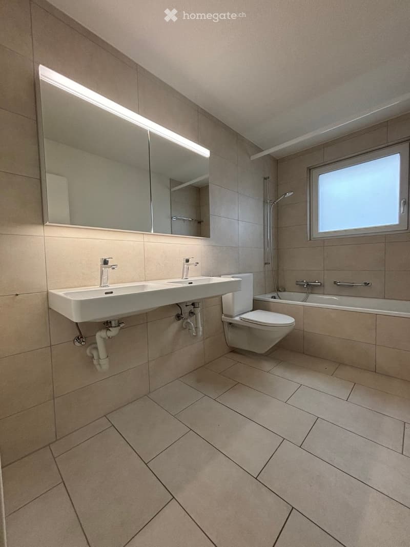 Modern bathroom with beige tiles, a double sink, toilet, bathtub, and large mirror with built-in lighting.