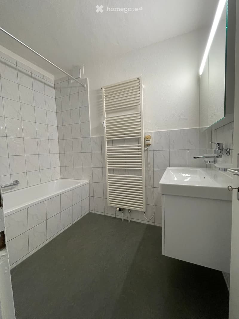 Modern bathroom with a bathtub, sink, radiator, and mirror. The walls are tiled, and the floor is dark grey.