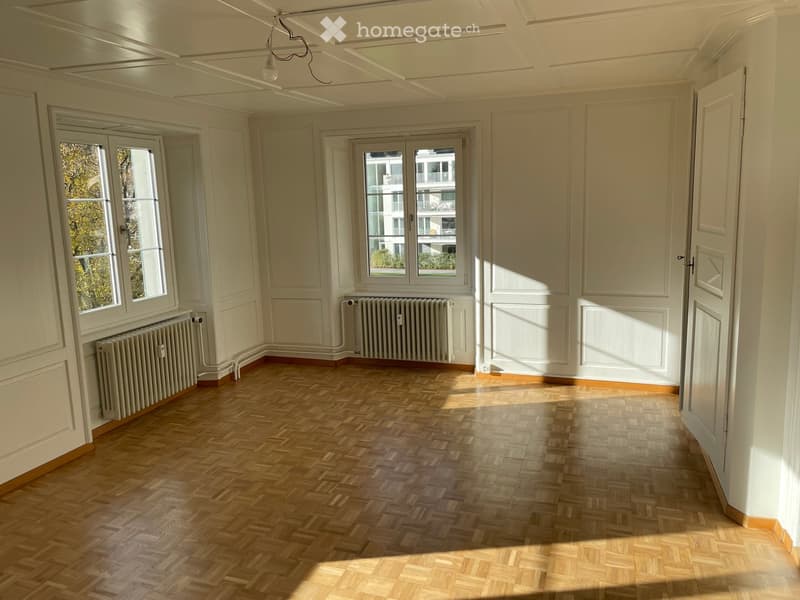 Empty room with wooden herringbone flooring, white walls, large windows, radiators, and a ceiling light fixture.