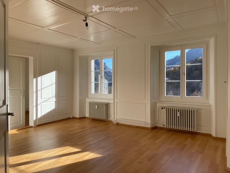 Empty room with wooden flooring, white walls, large windows offering a scenic mountain view, radiators, and a door leading to another space.