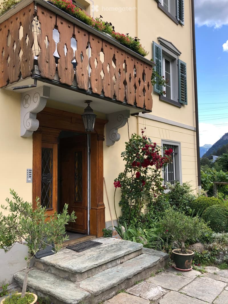 Front entrance of a house with a wooden balcony, stone steps, and garden plants.