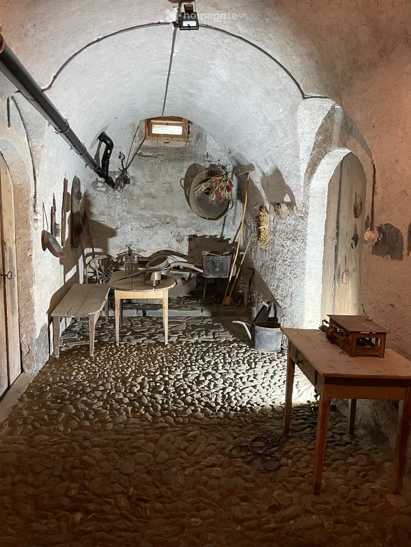 A rustic, stone-walled room with a stone floor, wooden tables, benches, and various tools and household items.