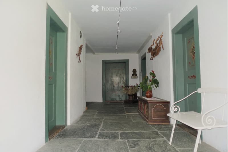 A long, narrow hallway with white walls, green doors, and a stone-tiled floor. Features include a small wooden bench with a potted plant, a white metal chair, and decorative wall art.