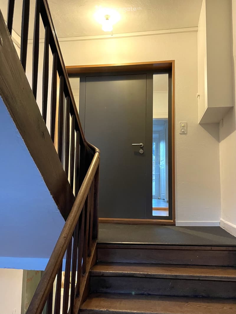 Wooden staircase with metal railing, closed door at the end, light fixture on the wall, and a small shelf above the door.