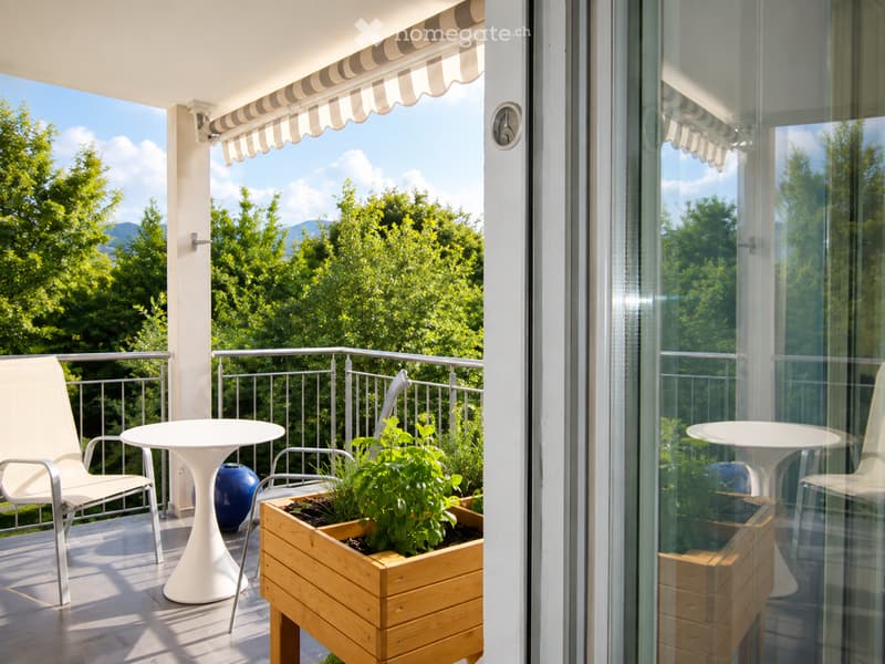 Balcony with a view of green trees, featuring a white chair, a white table, and a wooden planter with plants.