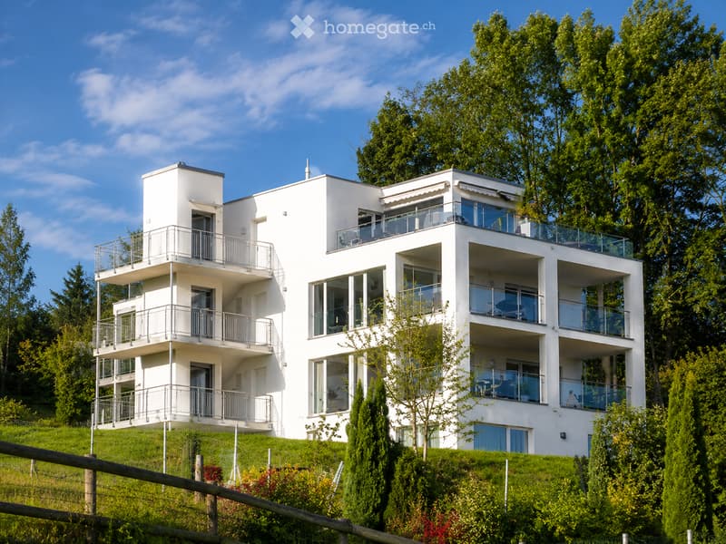 Modern white multi-story building with multiple balconies and large windows, surrounded by greenery and trees.