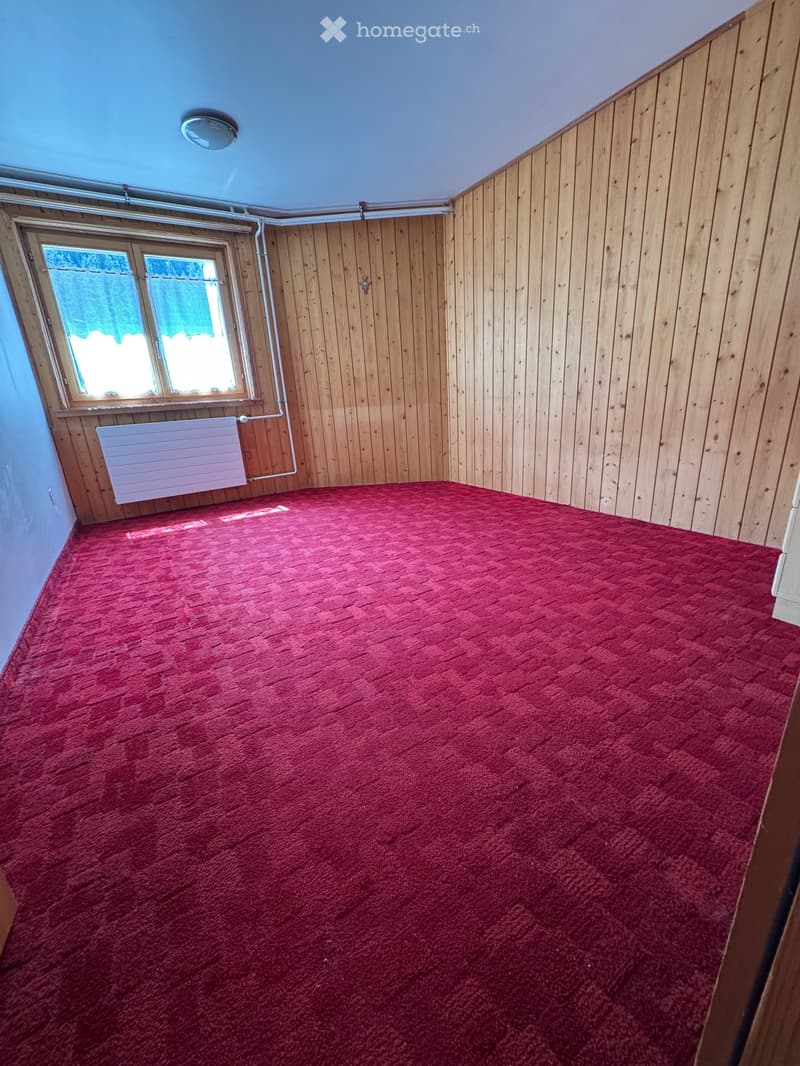 Empty room with red carpet, wooden walls, and a single window with blinds. No furniture or equipment present.