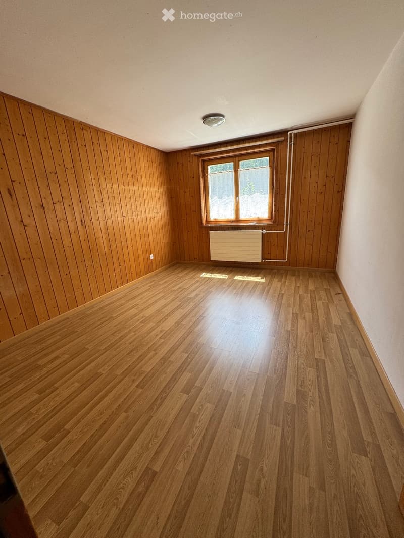 Empty room with wooden walls and floor, a window, radiator, and ceiling light fixture.