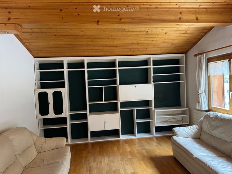 Living room with light wooden flooring, white built-in bookshelf with multiple compartments and cabinets, two beige sofas, and a window with white curtains.