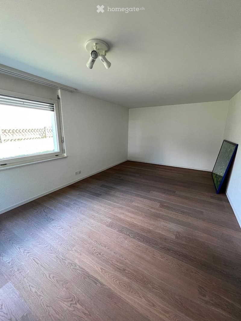 Empty room with wooden flooring, white walls, and a ceiling light fixture. A window with blinds is on one wall, and a mirror leans against another wall.