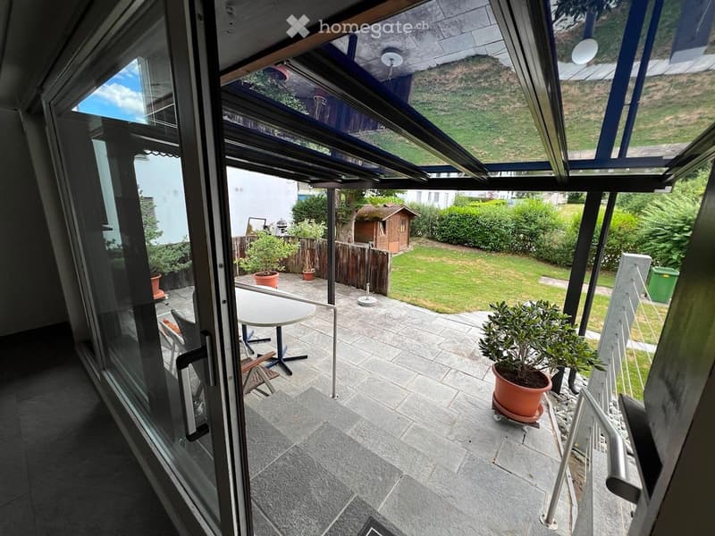 Glass-enclosed balcony with a table, chairs, and potted plants, overlooking a garden with a small wooden shed and lawn.