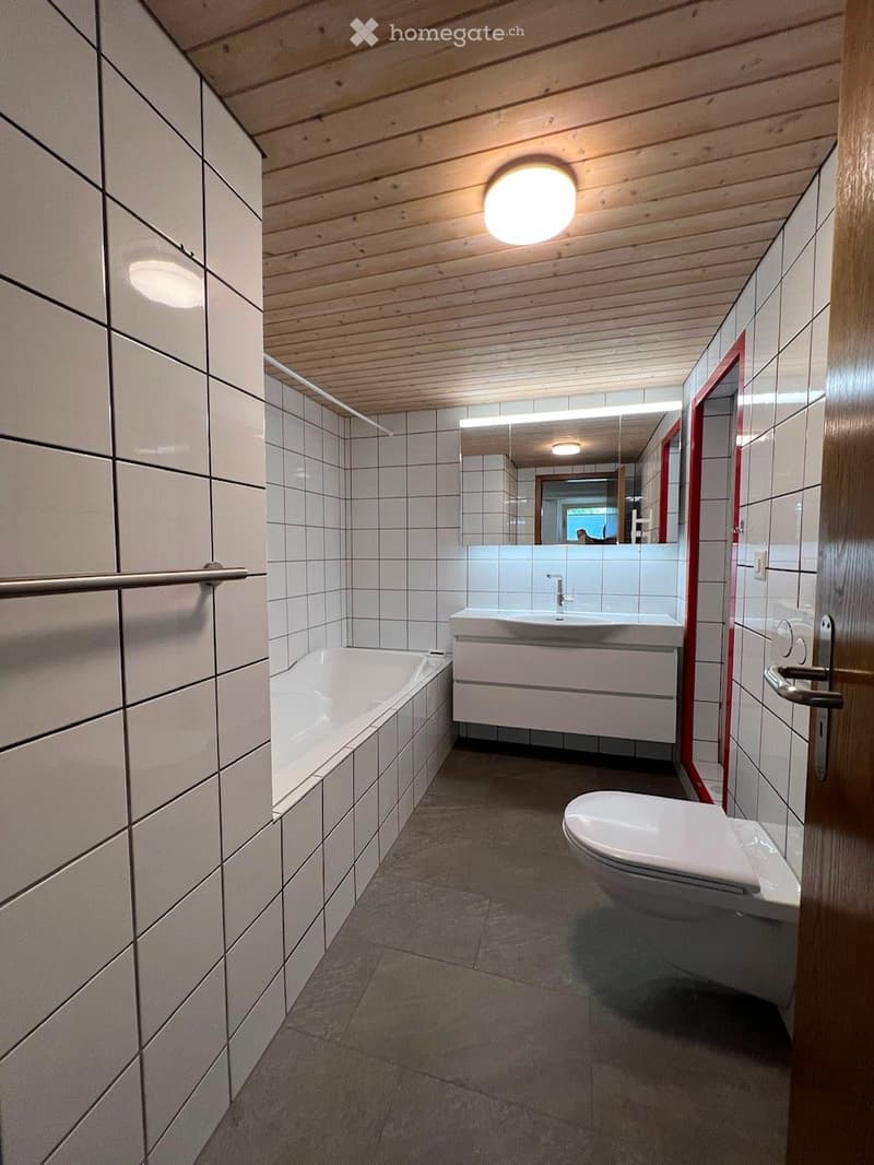 Modern bathroom with white tiled walls, a bathtub, toilet, sink, and a large mirror above the sink. The ceiling is wooden with recessed lighting.