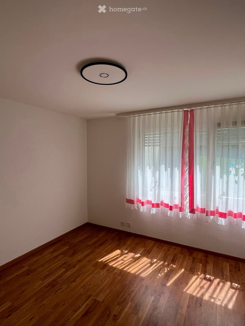 Empty room with wooden flooring, light pink walls, and white curtains with red trim on the windows. A round ceiling light fixture is visible.