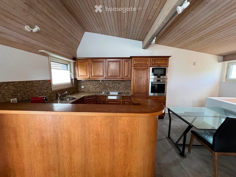 A modern kitchen with wooden cabinets, granite countertops, stainless steel appliances, and a bar counter. A glass dining table with a black leather chair is visible.