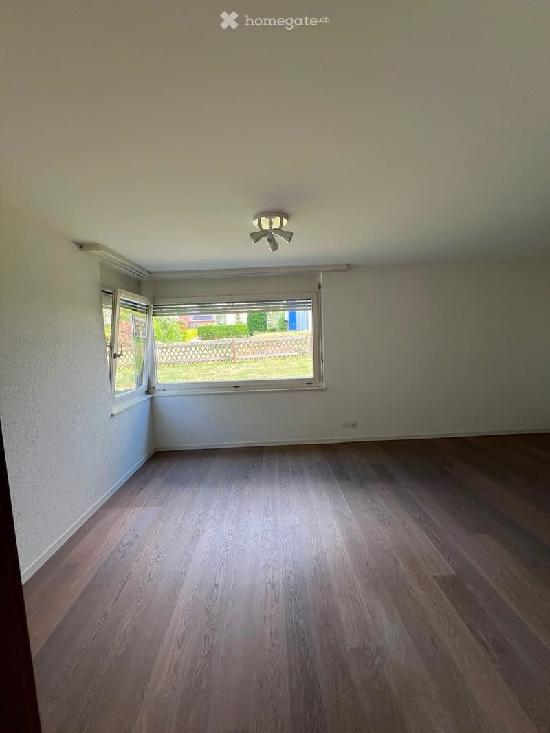Empty room with wooden flooring, white walls, and large windows offering a view of a garden area.
