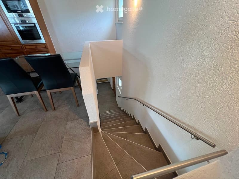 A modern staircase with beige tiled steps and a metal handrail, adjacent to a dining area with two black chairs and a glass table.